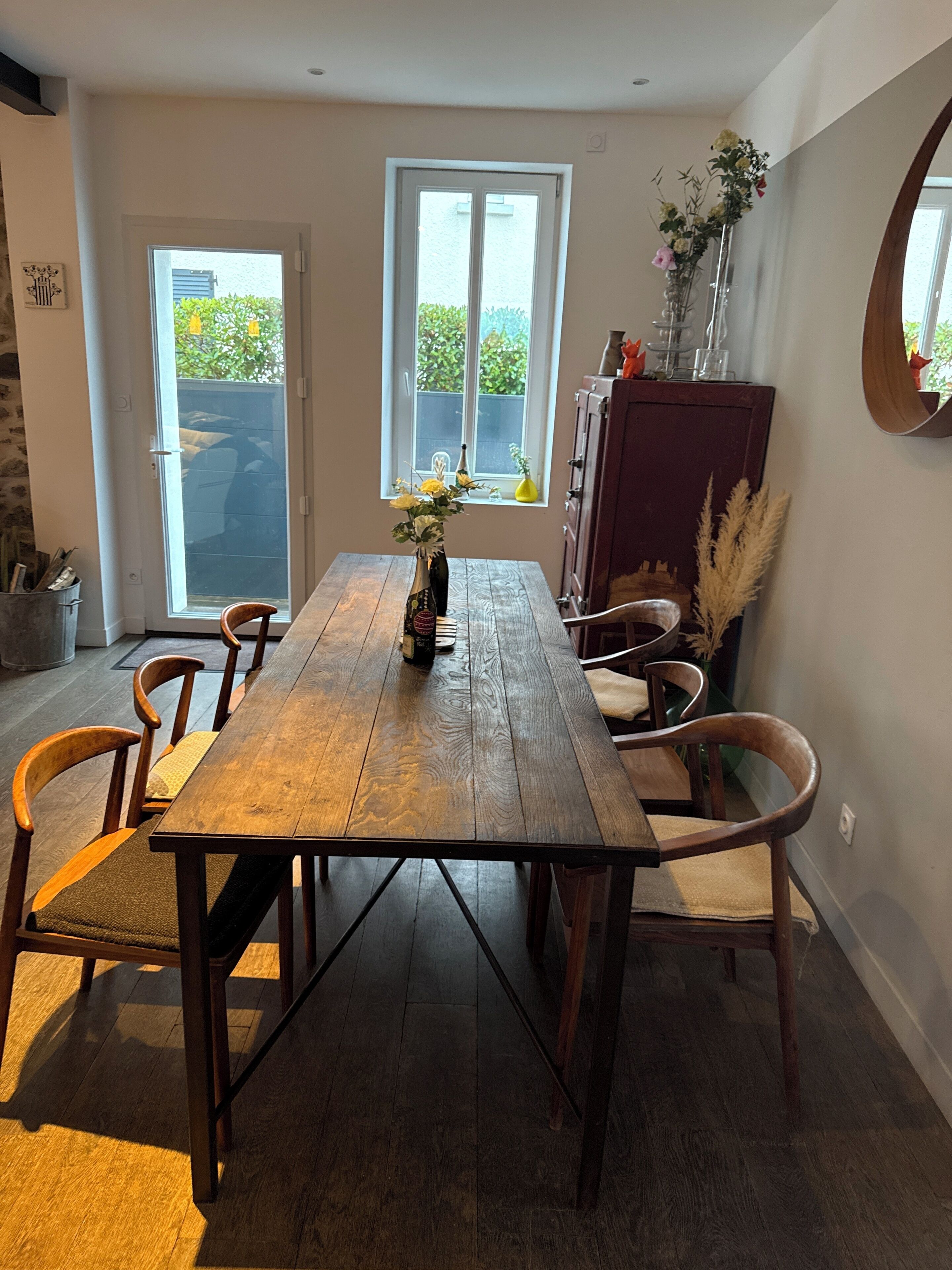 A dining area with a wooden table, chairs, a vase with flowers, and a view through a window.