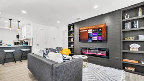 Ein modernes Wohnzimmer mit einem grauen Sofa, einem flachbildfernseher an der Wand, einem kleinen runden Tisch und einer Küchenzeile im Hintergrund.