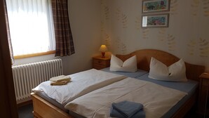 A double bed with two pillows, a bedside table with a lamp, a radiator, and a window with curtains. - Vacation home Mundt Between the Bodden and the Baltic Sea, at the very top in the north of Rügen (Altenkirchen OT Drewoldke)