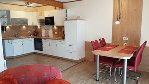 A compact kitchen with white cabinets, a stove, and a refrigerator. There is a dining table with red chairs and a red cushion. - Vacation home Mundt Between the Bodden and the Baltic Sea, at the very top in the north of Rügen (Altenkirchen OT Drewoldke)