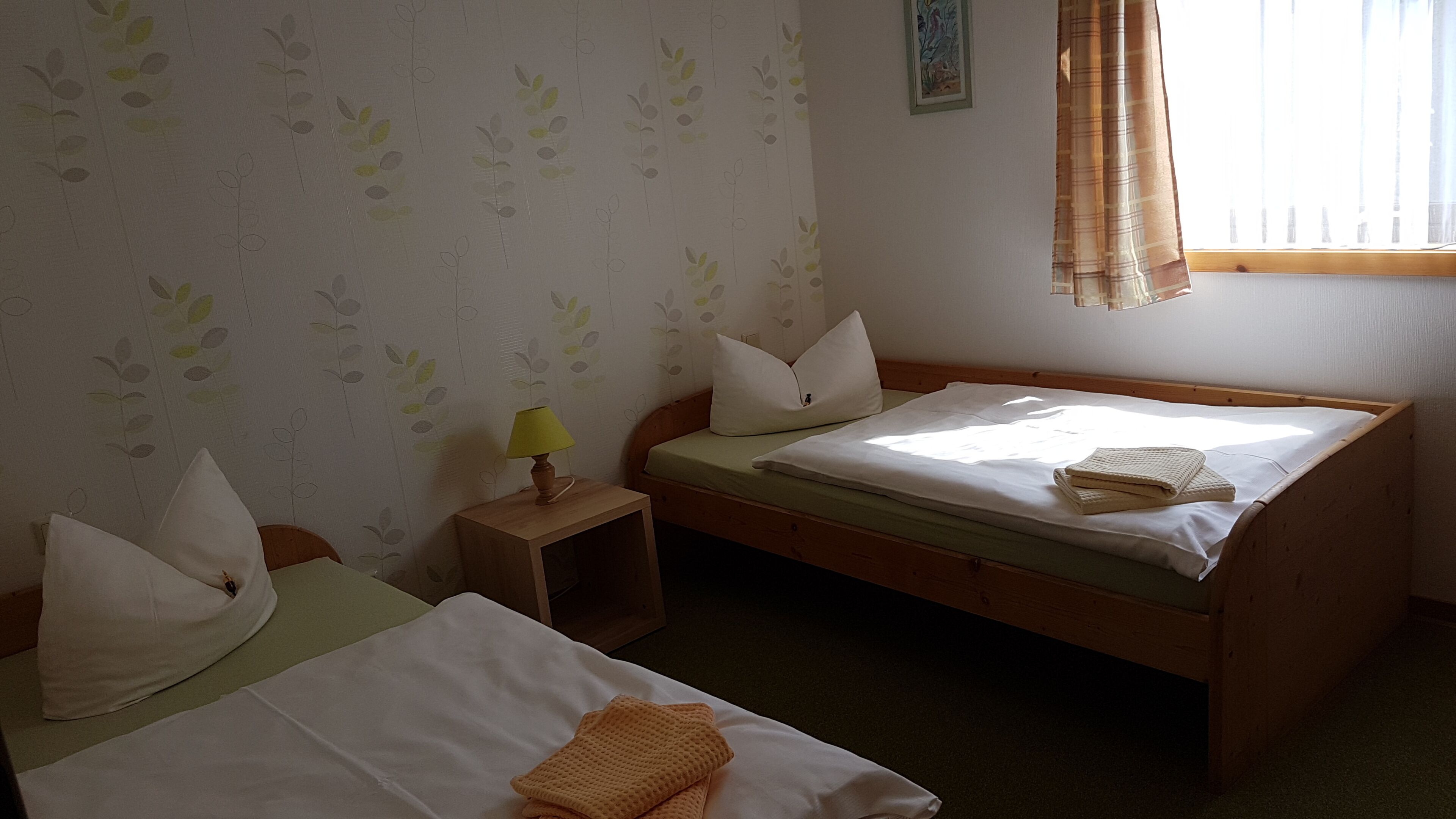 Ein schlichtes Zimmer mit zwei Betten, einem Nachttisch mit Lampe und einem Fenster mit Vorhängen.
