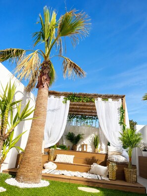 A cozy outdoor seating area with a thatched roof, white curtains, and a palm tree. - The love nest (Mèze)