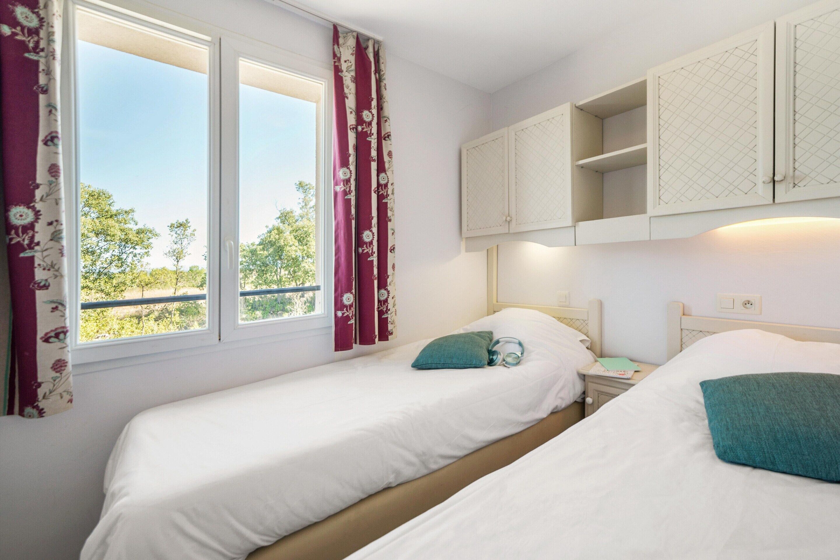 A bedroom with two beds, a window with curtains, a nightstand, and a cabinet above the beds.