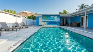 Ein Swimmingpool mit Wandgemälde einer Strandlandschaft, Liegestühlen und einer überdachten Sitzgruppe.