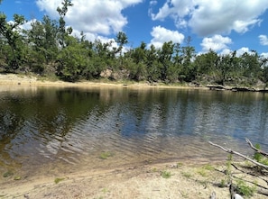 A calm lake with clear water, surrounded by trees and a sandy shore.