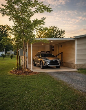 A covered garage with a parked SUV, a tree, and a house in the background.