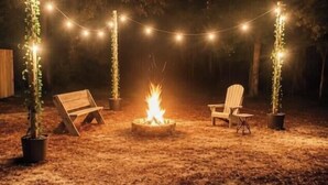 Een terras buiten met een vuurkorf, omringd door lichtslingers en potplanten.