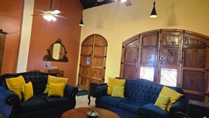 Una habitación con dos sofás azul oscuro, cojines amarillos, un mueble de madera, un ventilador de techo y una pequeña mesa redonda.
