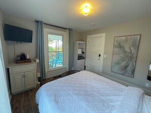 Un dormitorio con cama, televisor montado en la pared, ventana con cortinas, puerta y un cuadro en la pared.
