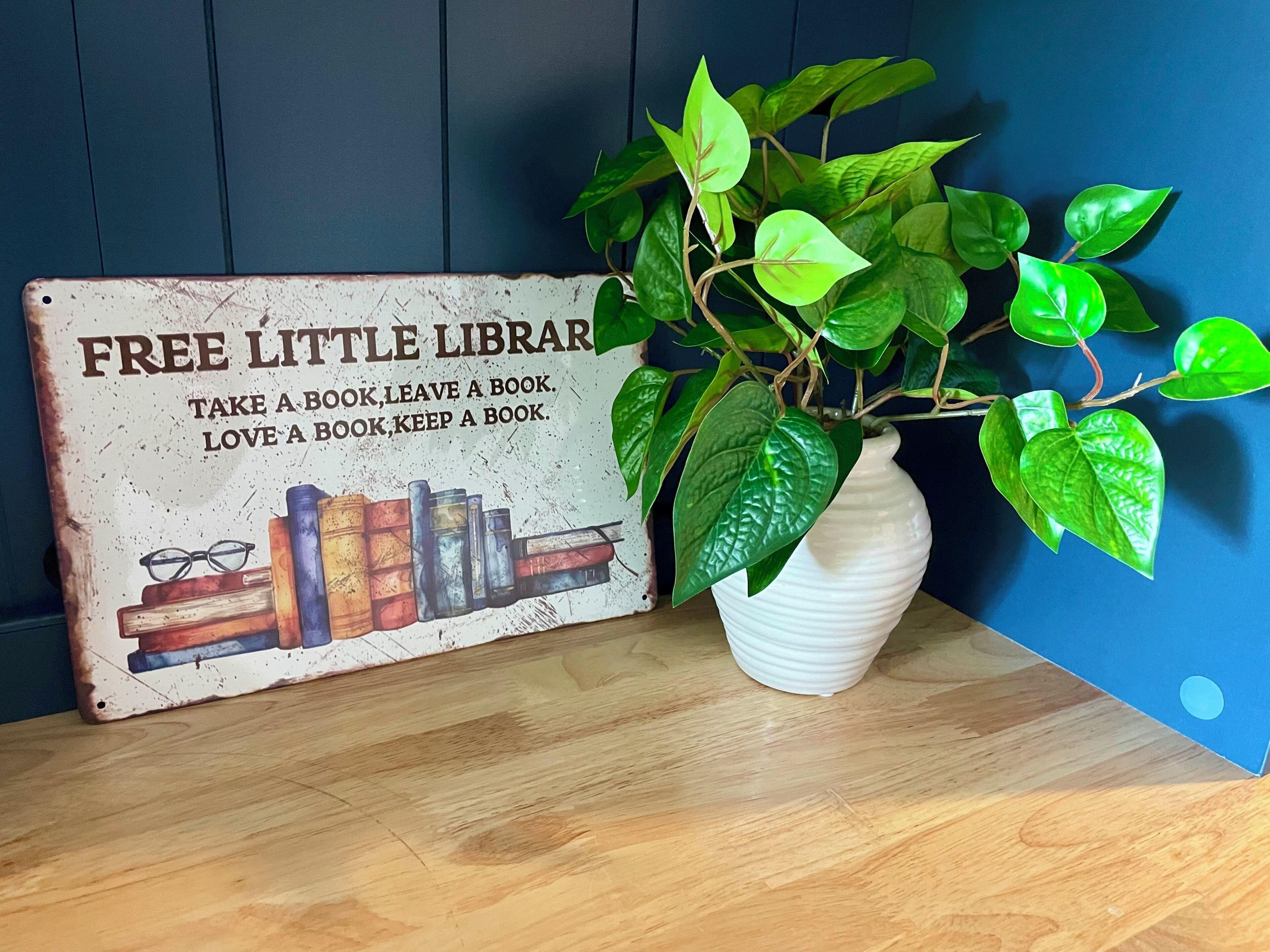 A sign with the text 'FREE LITTLE LIBRAR' and a potted plant beside it.