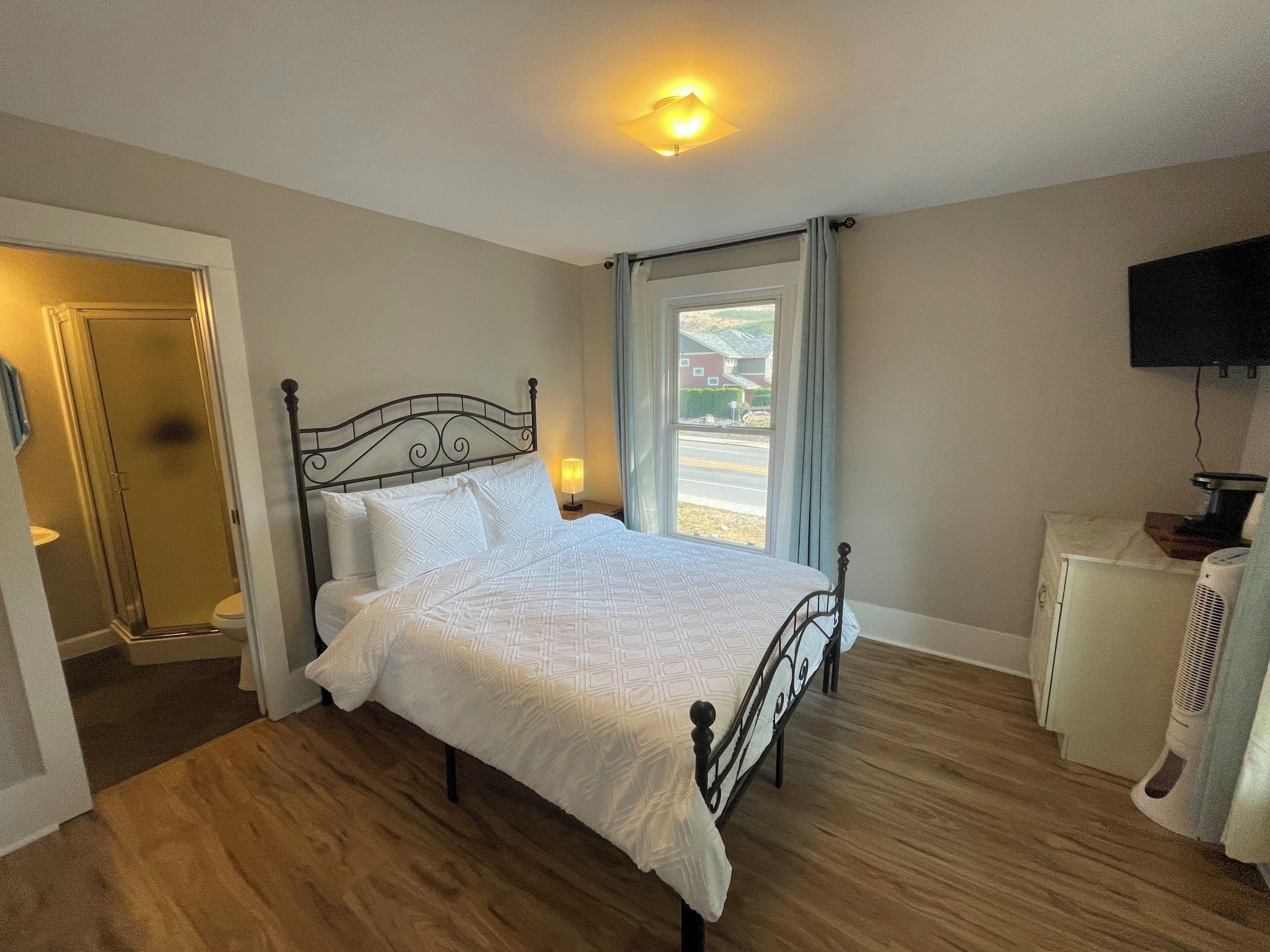 A bedroom with a bed, a bedside table, a lamp, a window with curtains, a television, and a heater.
