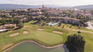 A golf course with a pond, surrounded by buildings with a prominent tower, and a clear sky. - Chic Riverfront Stay With Terrace (Mallemort)