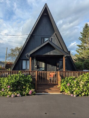 A triangular wooden cabin with a porch and a wooden fence.