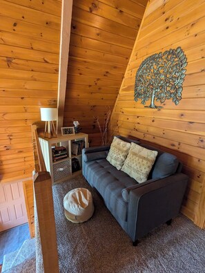 A cozy cabin interior with a gray sofa, a decorative metal tree wall art, a small white table, and a bookshelf.