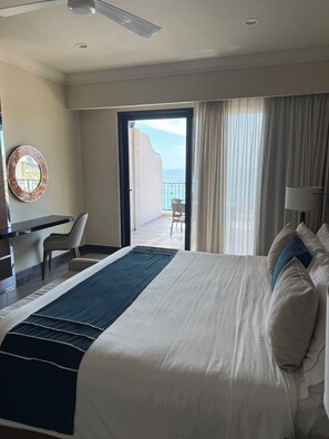 Habitación de hotel con cama, escritorio, silla, balcón con vista al océano y un espejo redondo en la pared.