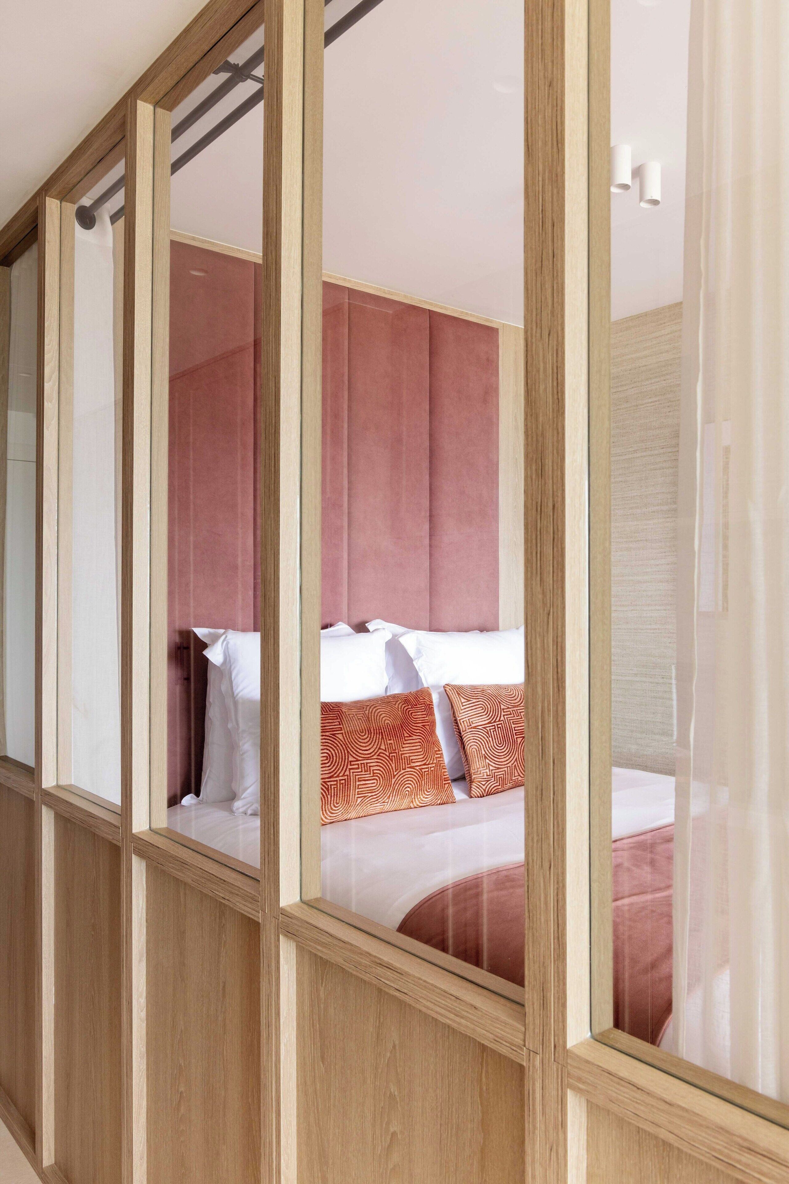 A modern hotel room with a large bed, wooden paneling, and a view through a glass partition.