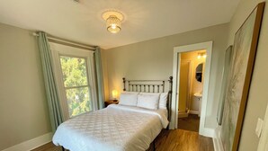 A bedroom with a bed, a nightstand, a lamp, a window with curtains, a door leading to a bathroom, and a ceiling light fixture. - Walk to Lake & Downtown Chelan! Room #5 (Chelan)