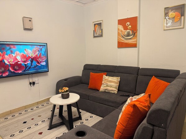 A modern living room with a grey sofa, orange cushions, a small round table with a vase, and a flat-screen TV on the wall.