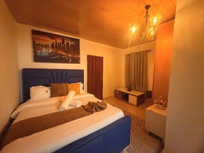 A hotel room with a bed, a desk, a chair, a chandelier, and a cityscape painting on the wall.