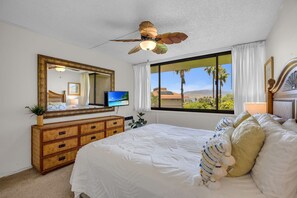 Ein Schlafzimmer mit Bett, Kommode, Fernseher, Deckenventilator und einem Fenster mit Blick auf Palmen.