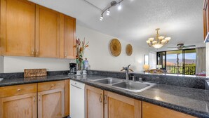 A kitchen with wooden cabinets, a granite countertop, a dishwasher, and a view of the outdoors. - KS4301 Partial Ocean Views, Two King Bedrooms, & Prime Kihei Location (Kihei)