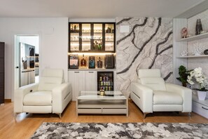 Ein modernes Wohnzimmer mit zwei weißen Ledersesseln, einem Glastisch, einer Wand mit Marmoroptik und einer integrierten Bar.
