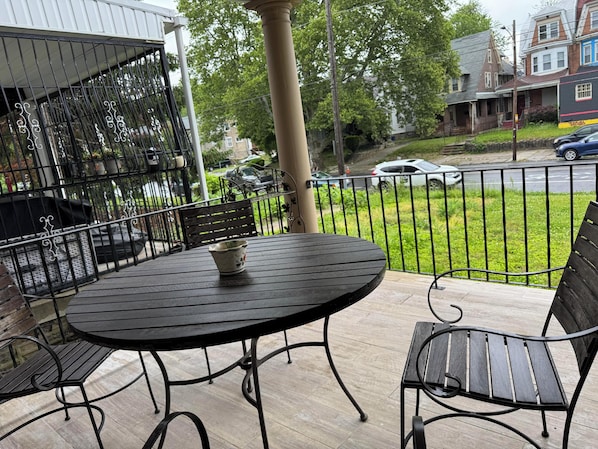 Un patio con una mesa redonda y dos sillas, baranda de metal, y vistas a la calle y casas.