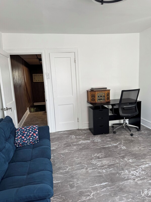A room with a blue sofa, a wooden desk with a chair, and a door leading to a hallway.