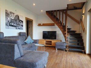 Ein modernes Wohnzimmer mit einem grauen Sofa, einem kleinen runden Tisch, einem Flachbildfernseher, Holzböden und einer Holztreppe.