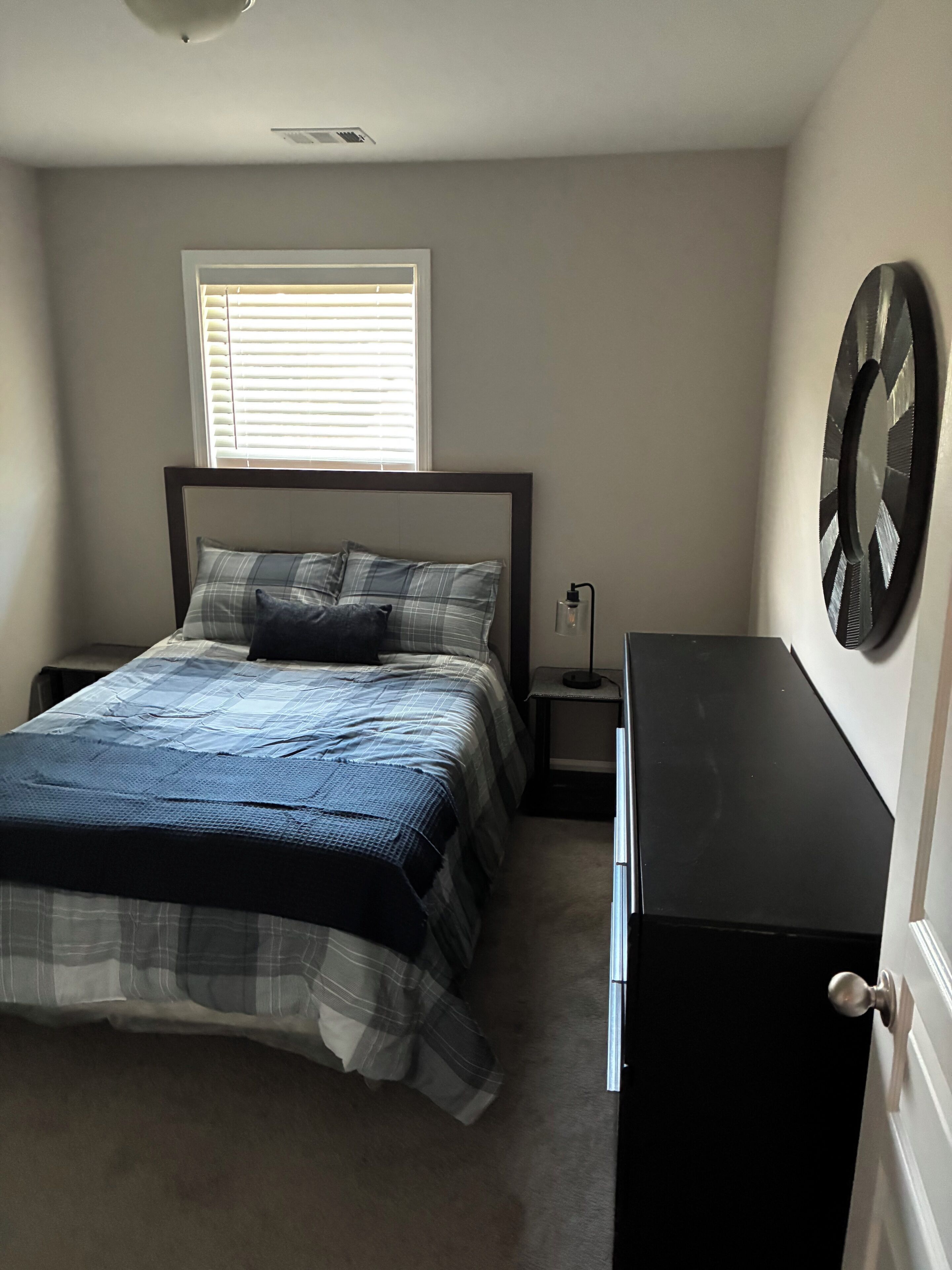 A bedroom with a bed, a dresser, a nightstand, a lamp, a window with blinds, and a wall clock.