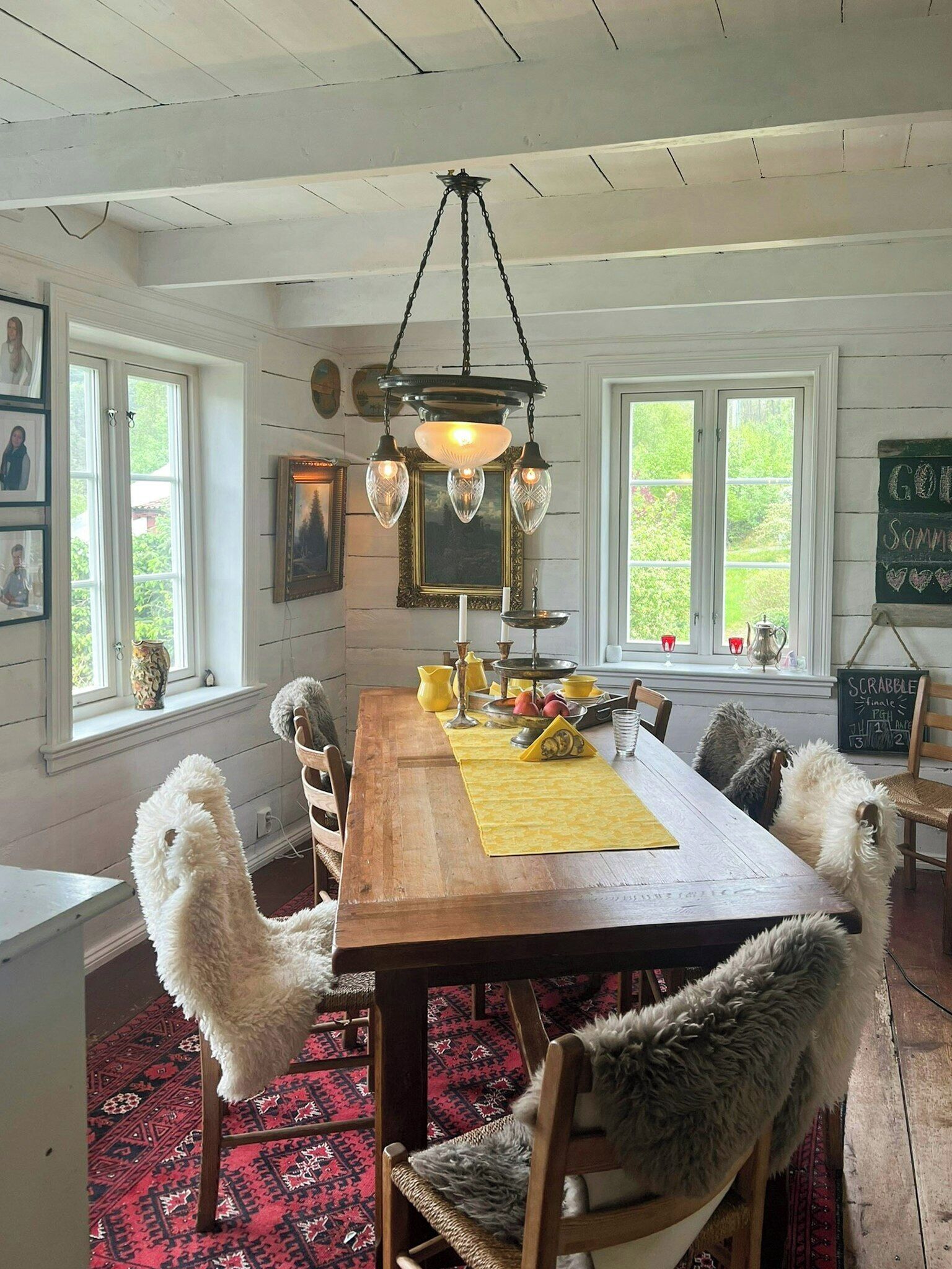 Une salle à manger avec une table en bois, des chaises, un lustre et une vue sur la verdure à l’extérieur.