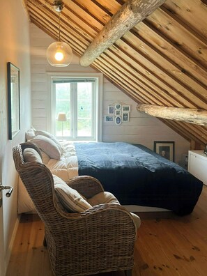 Une chambre avec un plafond en pente, un lit, une chaise en osier, une fenêtre et des tableaux accrochés au mur.