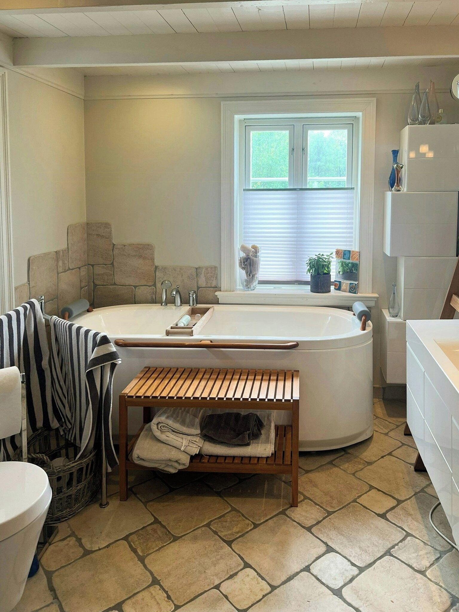 A bathroom with a freestanding bathtub, a wooden bench, a window with blinds, and a stone-tiled floor.