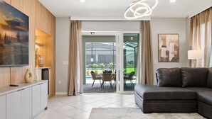 Un salon moderne comprenant un canapé d’angle, un téléviseur à écran plat, un coin repas avec des chaises et une porte vitrée donnant sur une terrasse extérieure.