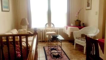A living room with a sofa, a dining table, a chair, a radiator, and a window with curtains.