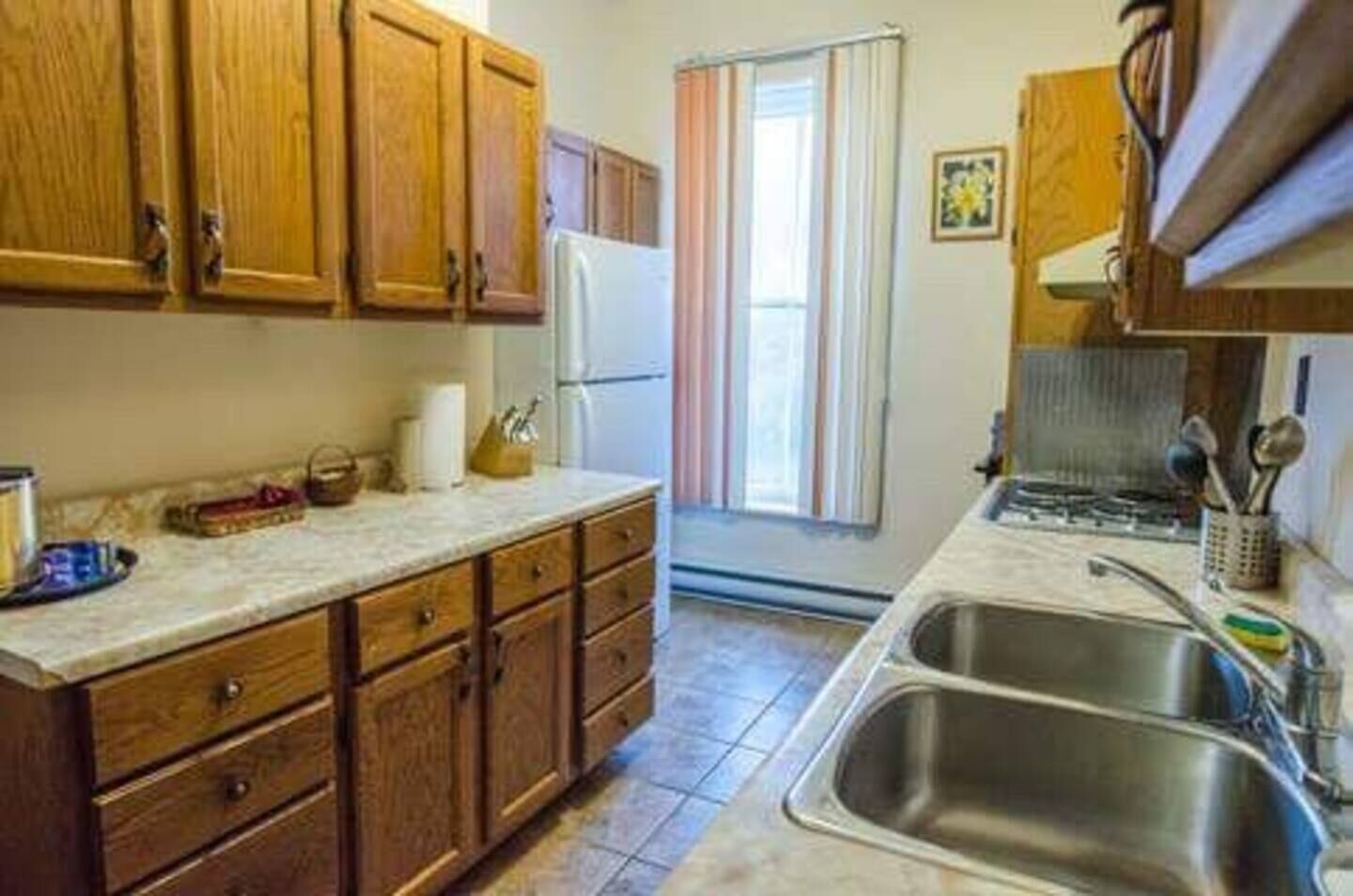 A kitchen with wooden cabinets, a refrigerator, a sink, and a window with curtains.