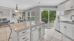 A modern kitchen with white cabinetry, granite countertops, and a large island. There is a dining area with a table and chairs, and a view of greenery through the sliding glass doors.