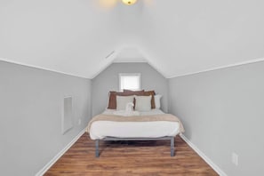 Un dormitorio con techo inclinado, una cama con ropa de cama blanca, una ventana y una rejilla de ventilación.