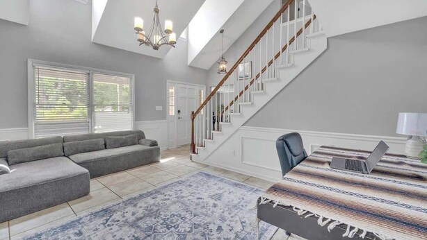 A spacious living room with a large gray sectional sofa, a patterned rug, a staircase with white railings, and a chandelier.