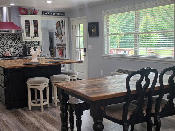 Una cocina con un letrero rústico, una isla de madera con taburetes, una mesa de comedor con sillas y una ventana con persianas.