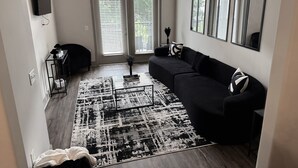 Una sala de estar con un sofá negro, una mesa de centro, un televisor montado en la pared y un ventanal con persianas.