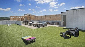 Un patio al aire libre con mobiliario y una mesa, una zona verde y un edificio con fachada de hormigón.