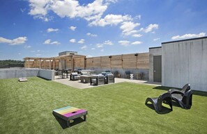 Un patio al aire libre con mobiliario y una mesa, una zona verde y un edificio con fachada de hormigón.