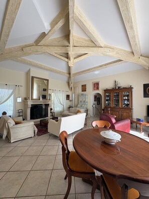 Ein großzügiges Wohnzimmer mit Kamin, Holzbalken, einem Esstisch mit Stühlen und einem roten Sofa.