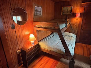 Ein Schlafraum mit Etagenbetten, Holzwänden, einem runden Spiegel, einem kleinen Tisch mit Lampe und zwei gerahmten Bildern.