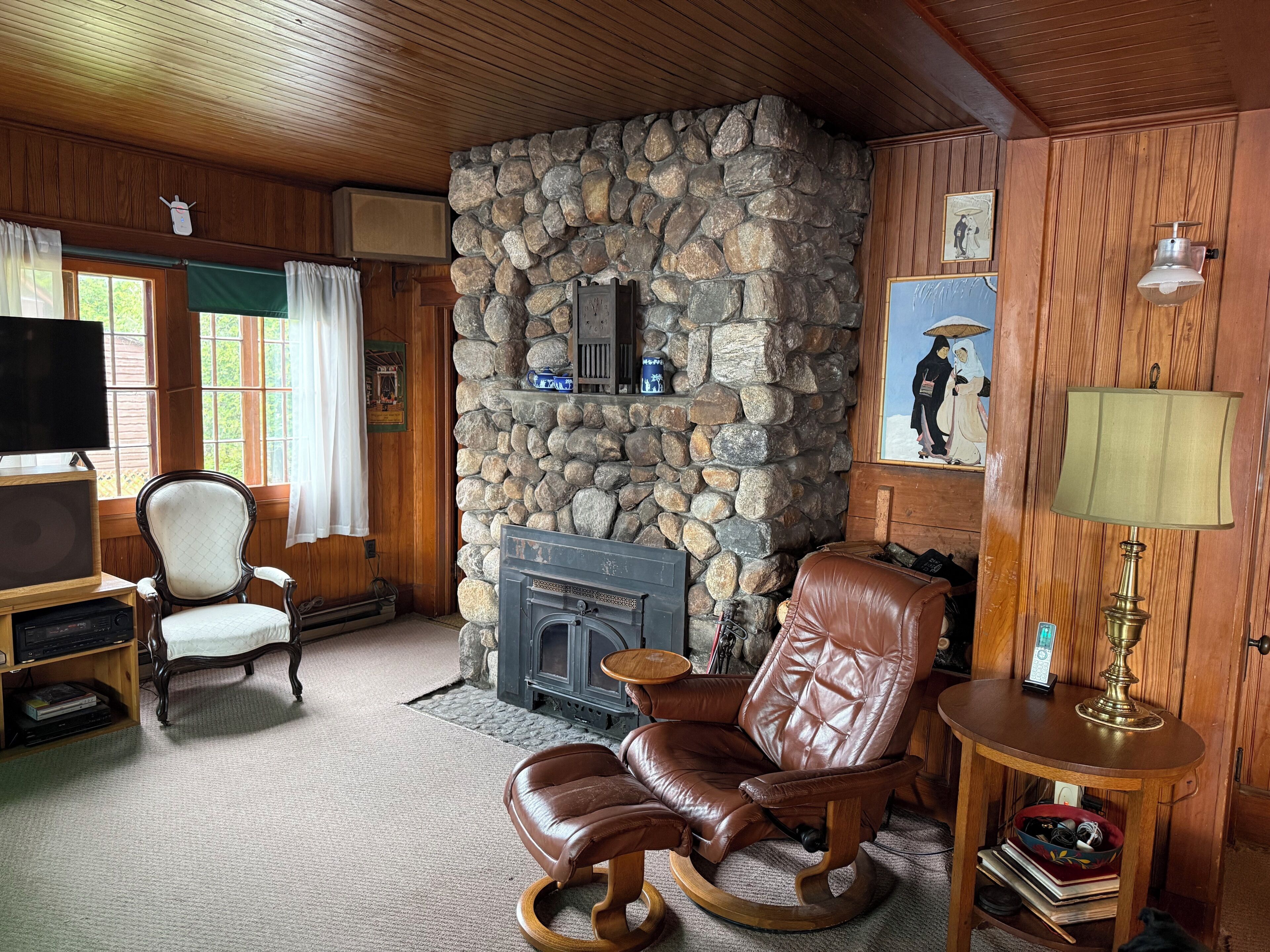 A cozy living room with a stone fireplace, wooden walls, a leather armchair, a television, and a small table with a lamp.