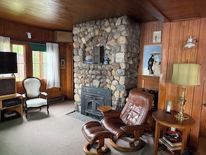 Ein gemütliches Wohnzimmer mit Kamin aus Stein, Holzwänden, einem Ledersessel, einem Fernseher und einem kleinen Tisch mit Lampe.