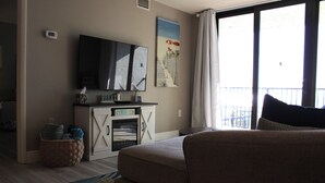 Een woonkamer met een flatscreen televisie, een schilderij, een bank, een open haard en uitzicht door een raam.