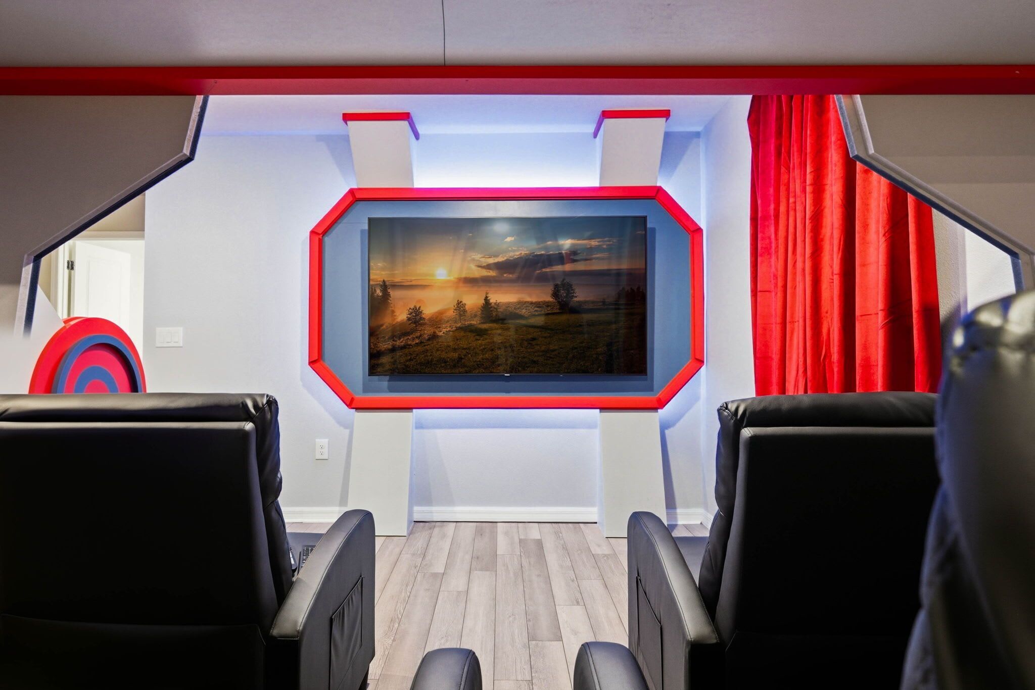 A room with a large TV mounted on a white wall, two black leather chairs, a red curtain, and a target on the wall.