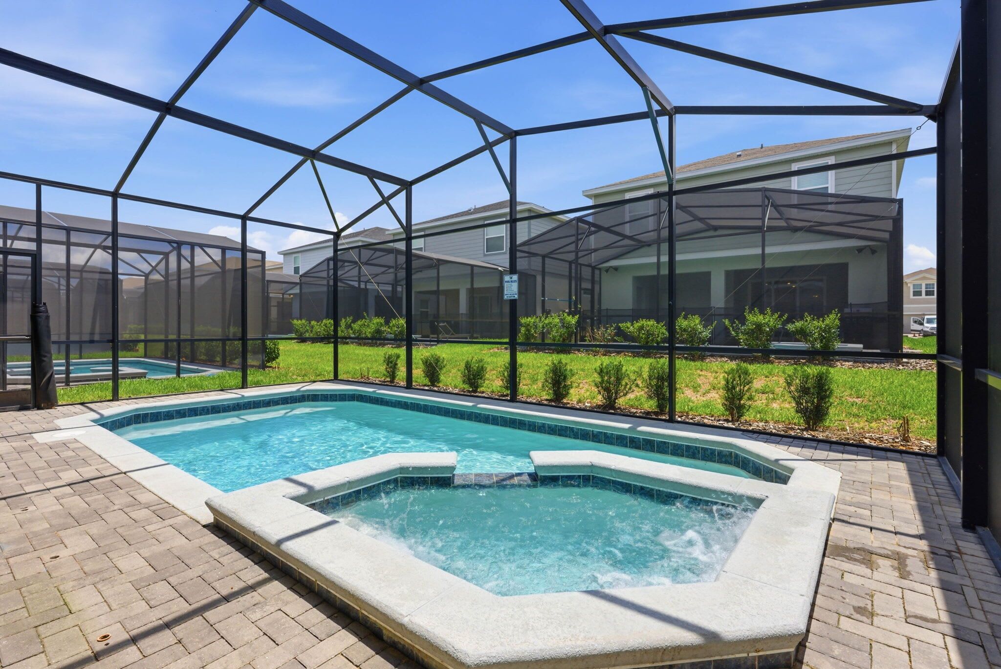 Zona de piscina con jacuzzi, rodeada por un recinto de vidrio y vistas a casas al fondo.